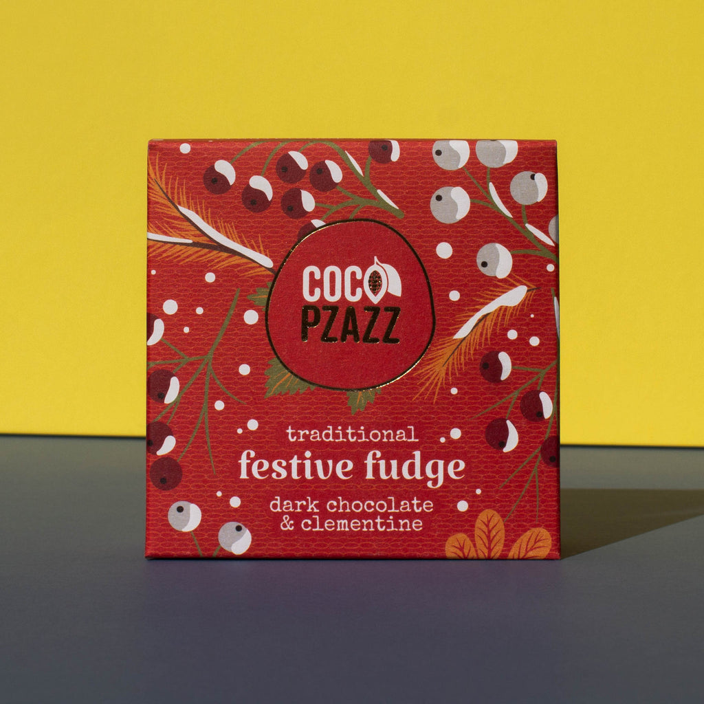 Box of Coco Pzazz Dark Chocolate and Clementine Fudge, it is a red box with an illustration of winter berries, it is placed on a blue and yellow background.