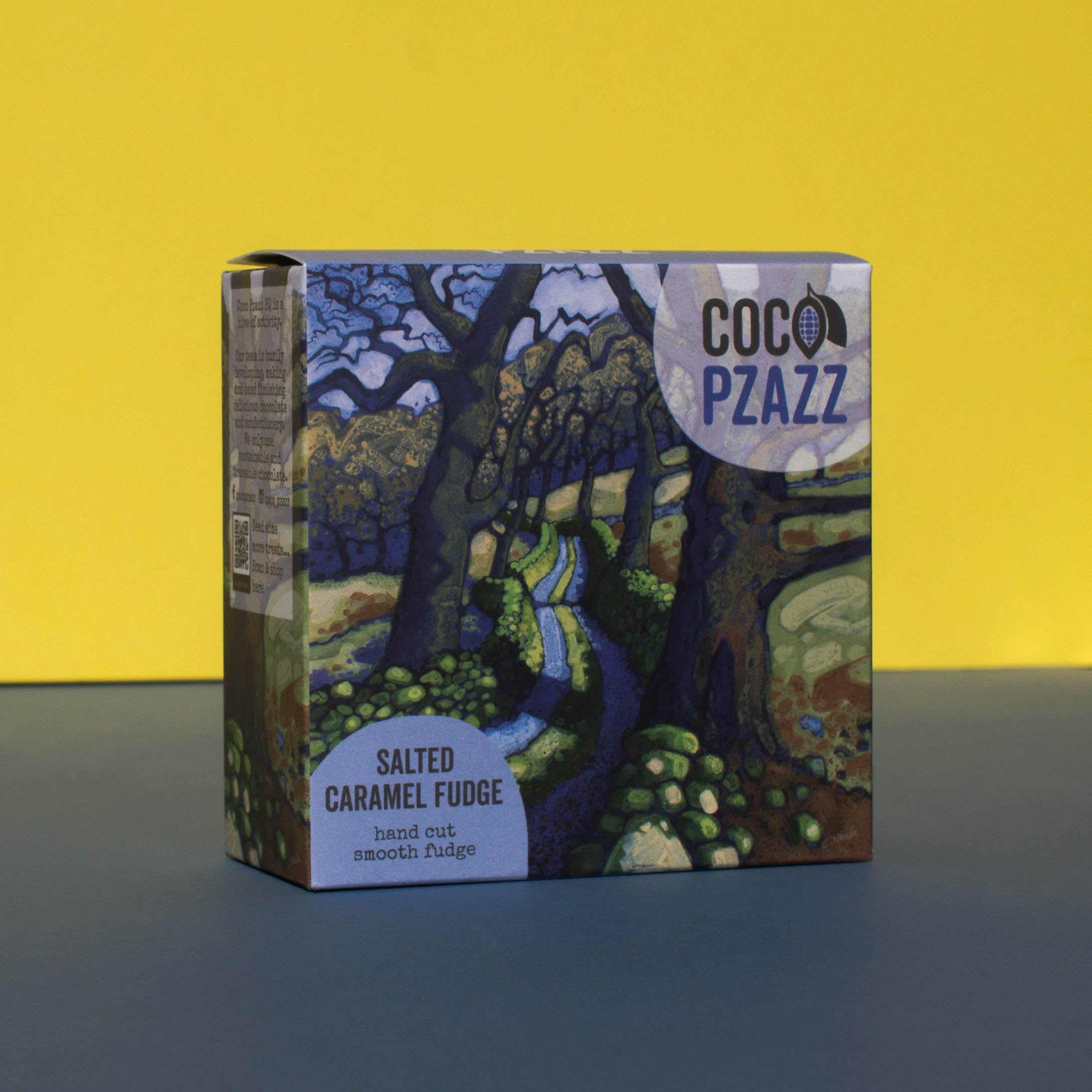 Box of Coco Pzazz Salted Caramel Fudge, it has an illustration of a country lane lined with trees, it is placed on a blue and yellow background and tilted slightly to the side.