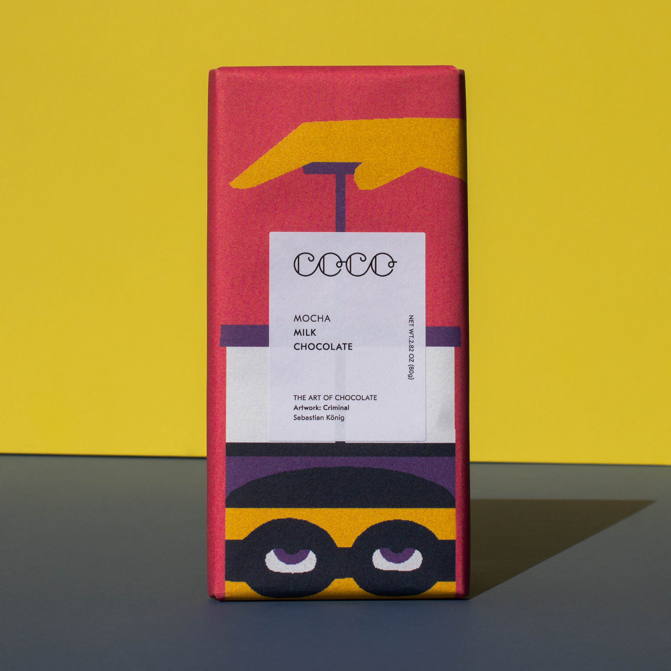 Bar of Coco Chocolatier Mocha Milk Chocolate, it has a pink wrapper with an illustration of a French press, it is placed on a blue and yellow background.
