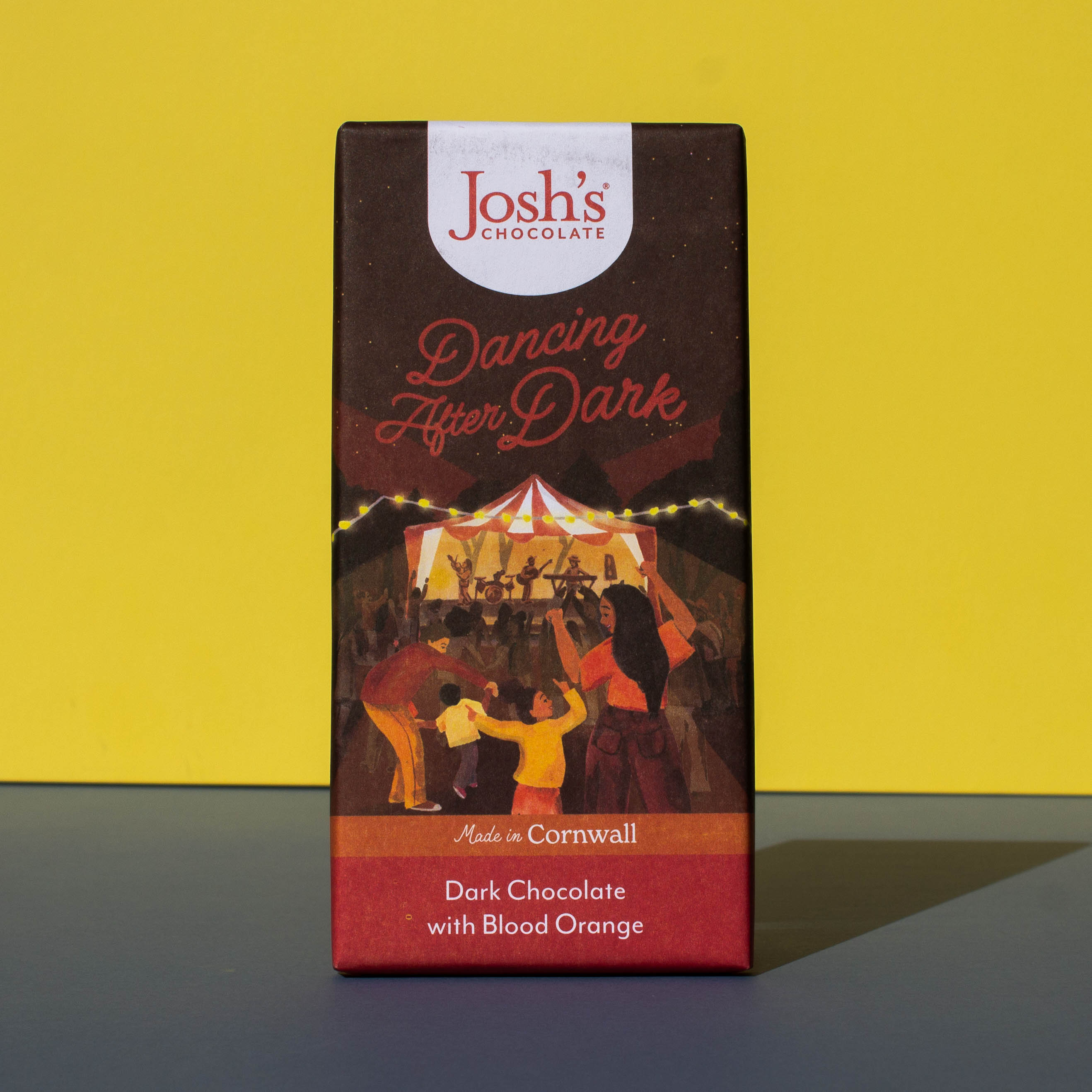 Bar of Josh's Dark Chocolate with Blood Orange, it is a dark orange and red paper wrapper with an illustration of an outdoor music event, it is placed on a blue and yellow background.