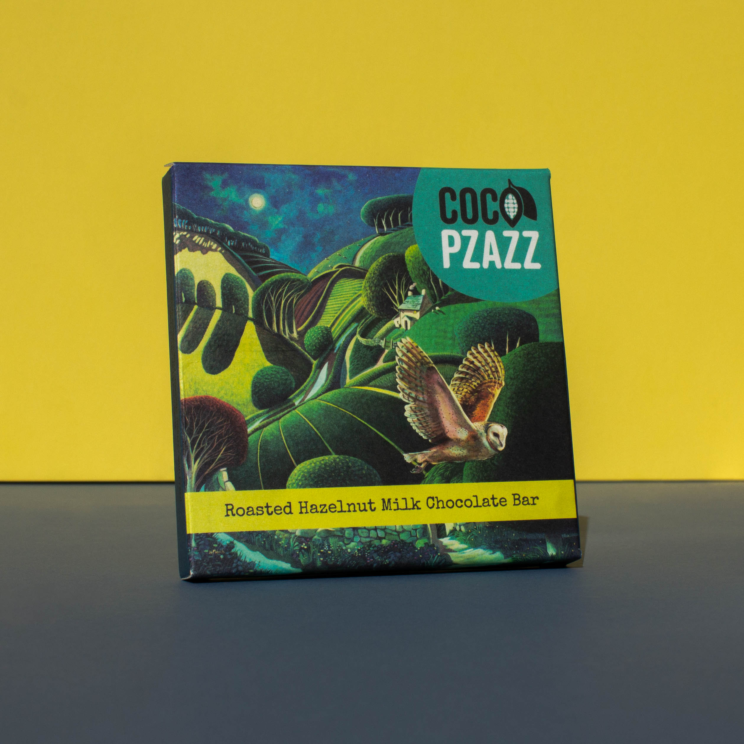 Bar of Coco Pzazz Roasted Hazelnut Milk chocolate, it has a dark blue wrapper with an illustration of an owl flying over farmland, it is placed on a blue and yellow background and tilted slightly to the side.