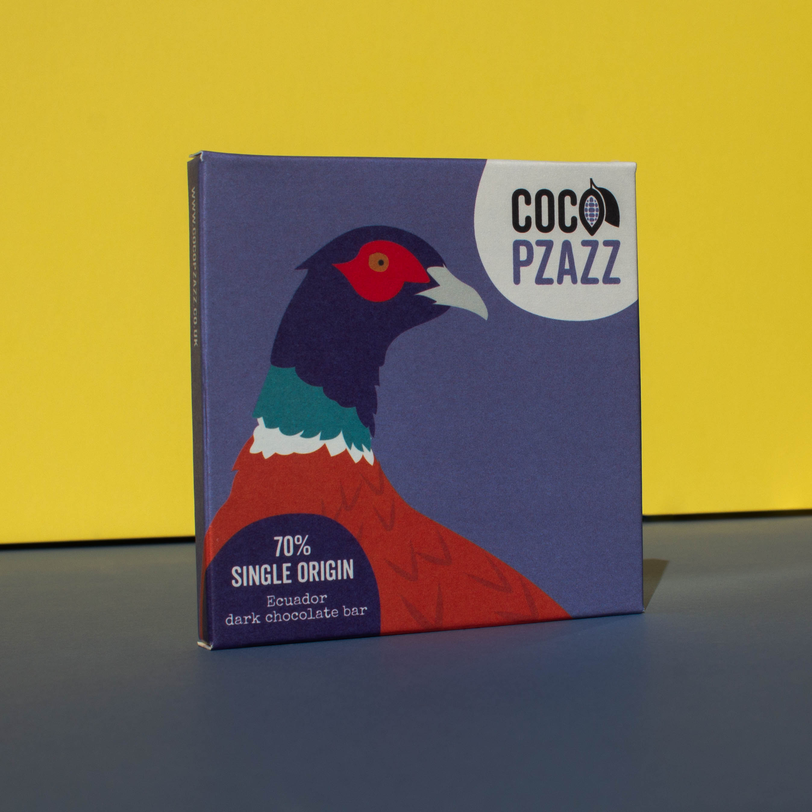 Bar of Coco Pzazz Ecuador Dark Chocolate, it has a blue wrapper with an illustration of a pheasant, it is placed on a blue and yellow background and is tilted slightly to the side.