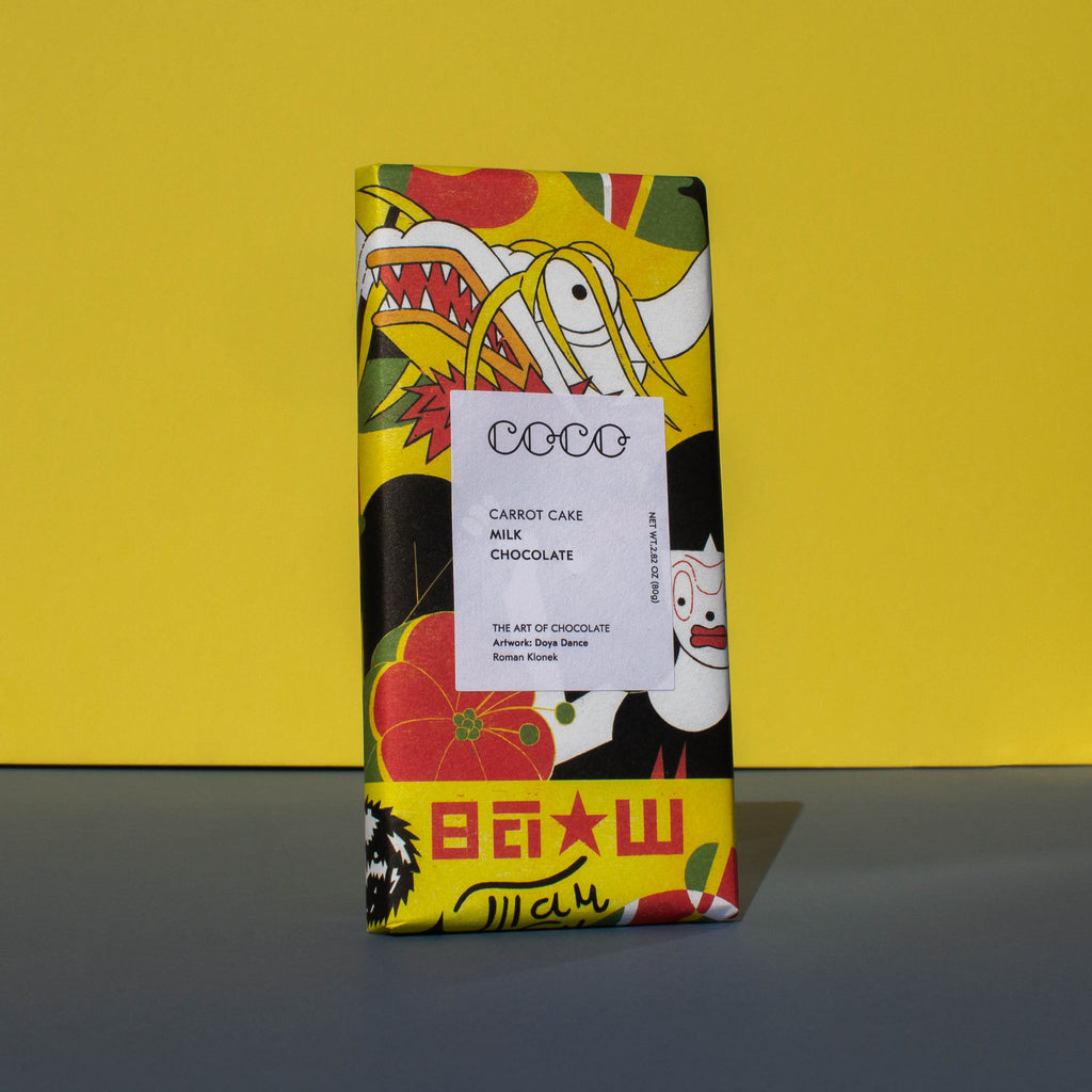 Bar of Coco Chocolatier Carrot Cake Milk Chocolate, it has a brightly colourful yellow, red and black illustration of a dragon on the wrapper, it is placed on a blue and yellow background and is tilted slightly to the side.
