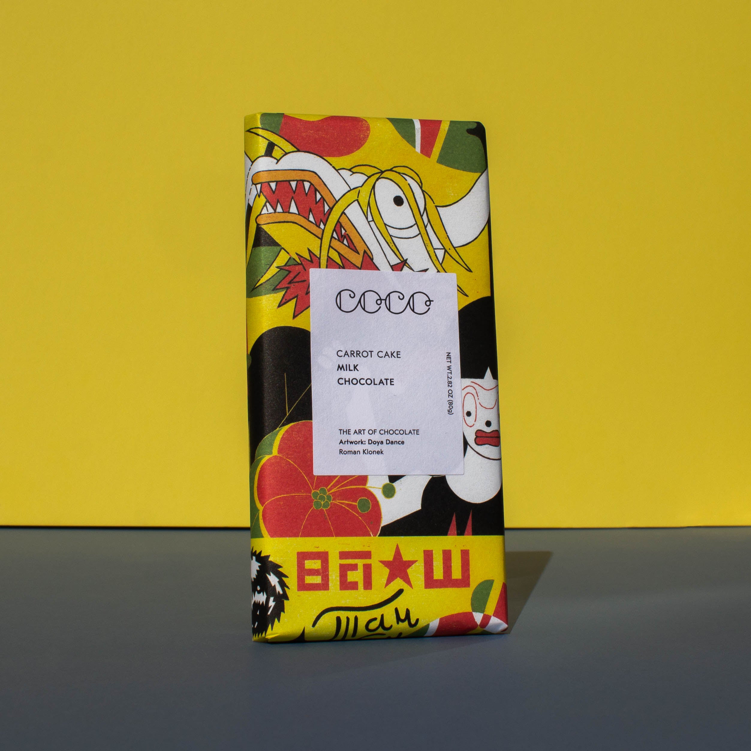 Bar of Coco Chocolatier Carrot Cake Milk Chocolate, it has a brightly colourful yellow, red and black illustration of a dragon on the wrapper, it is placed on a blue and yellow background and is tilted slightly to the side.