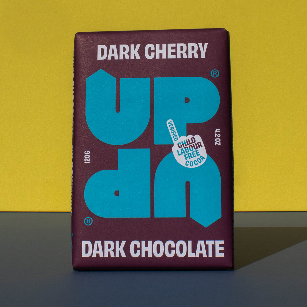 Bar of UP UP Dark Cherry Dark Chocolate, it has a purple paper wrapper with a blue logo, it is placed on a blue and yellow background.