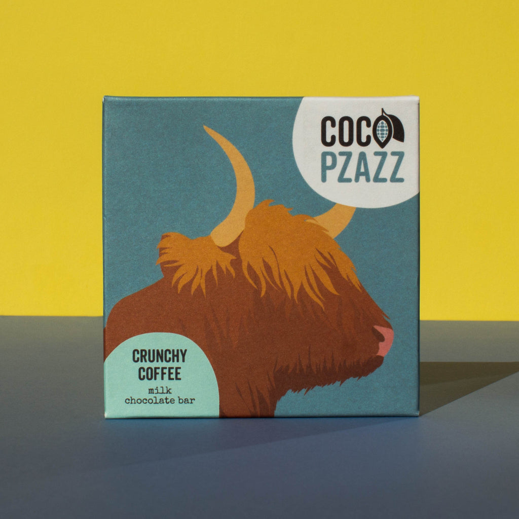 Bar of Coco Pzazz Crunchy Coffee Milk Chocolate, it has a blue wrapper with an illustration of a highland cow, it is placed on a blue and yellow background.