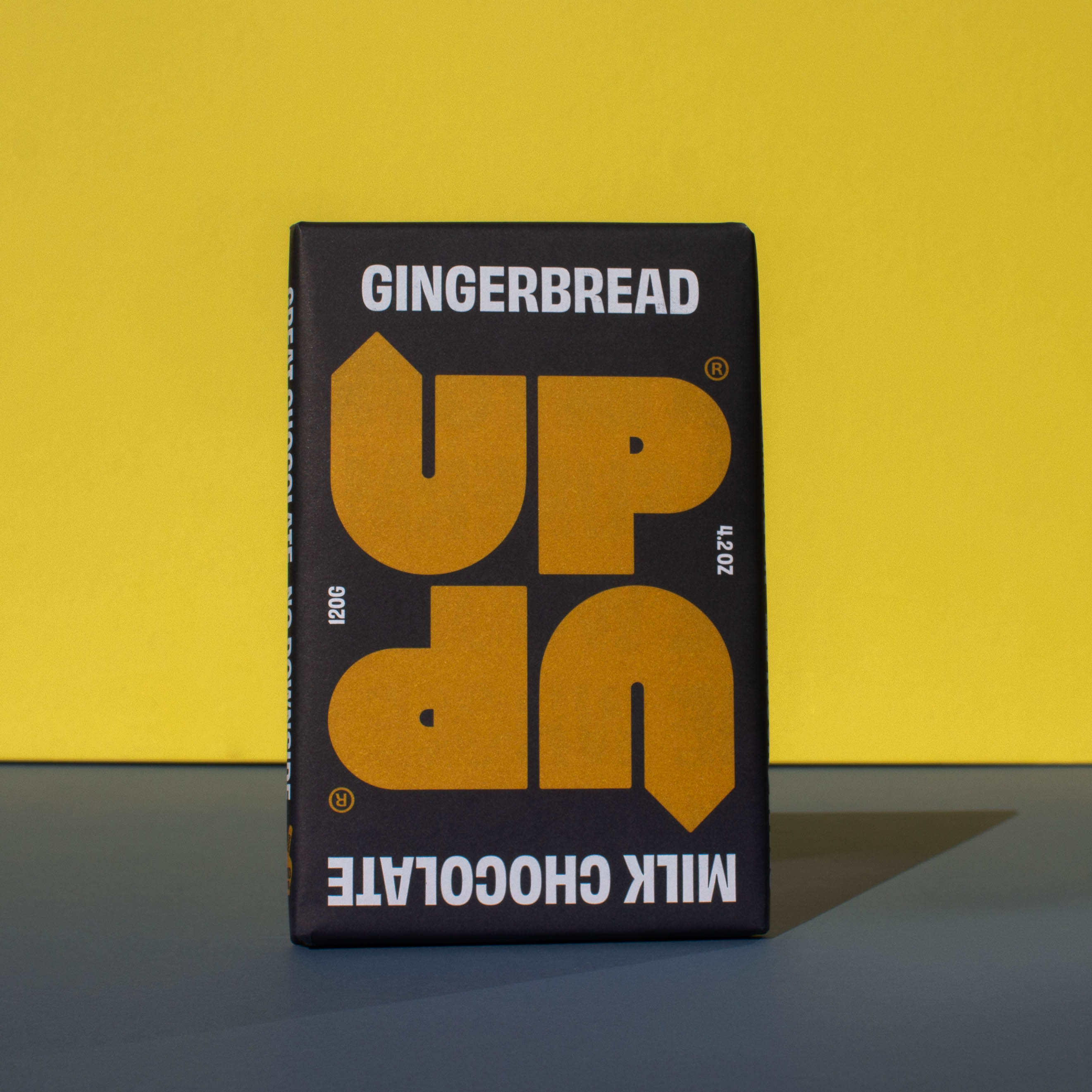 Bar of UP UP Gingerbread Milk Chocolate, it has a black paper wrapper with a yellow logo, it is placed on a blue and yellow background.