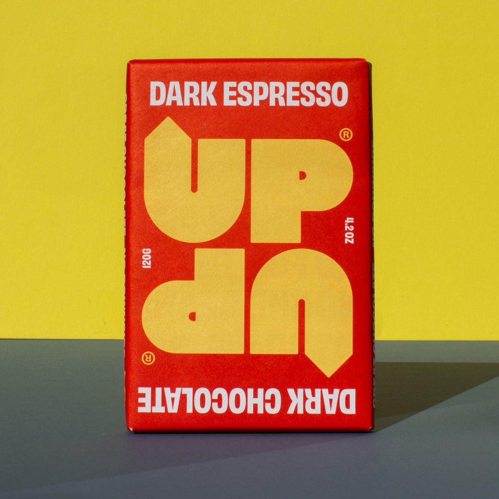 Bar of UP UP Dark Espresso Dark Chocolate, it has a red paper wrapper with a yellow logo, it is placed on a blue and yellow background.