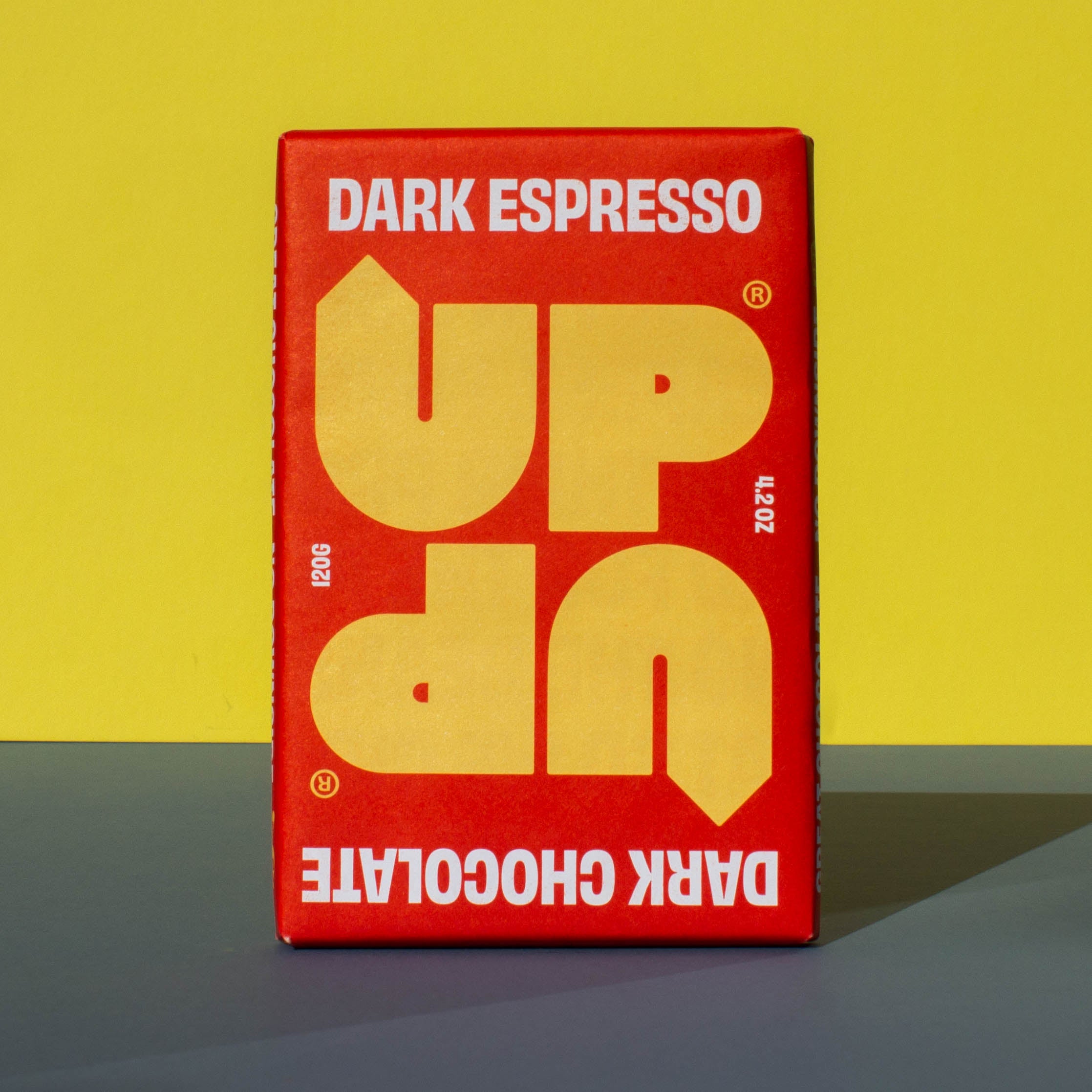 Bar of UP UP Dark Espresso Dark Chocolate, it has a red paper wrapper with a yellow logo, it is placed on a blue and yellow background.