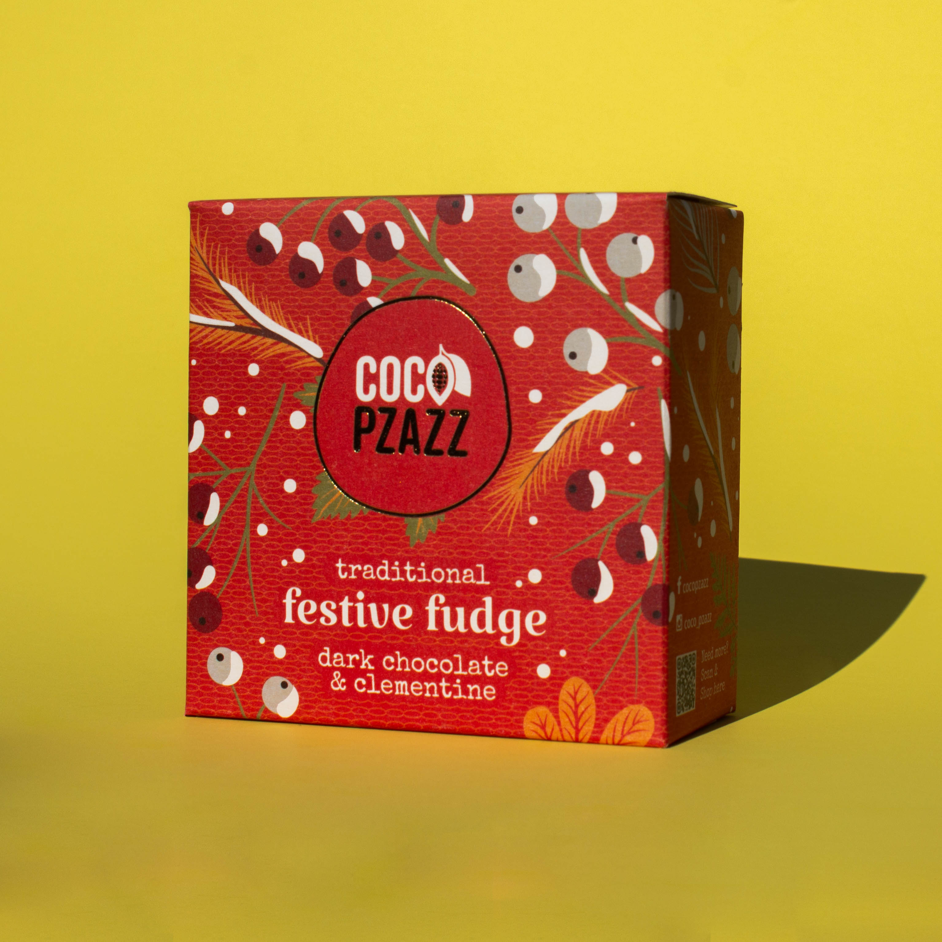 Box of Coco Pzazz Dark Chocolate and Clementine Fudge, it is a red box with an illustration of winter berries, it is placed on a yellow background and tilted slightly to the side.