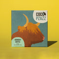 Bar of Coco Pzazz Crunchy Coffee Milk Chocolate, it has a blue wrapper with an illustration of a highland cow, it is placed on a yellow background.