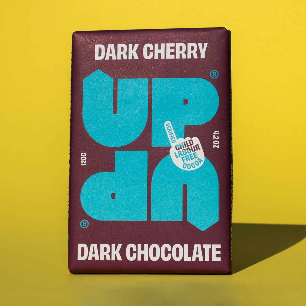 Bar of UP UP Dark Cherry Dark Chocolate, it has a purple paper wrapper with a blue logo, it is placed on a yellow background.