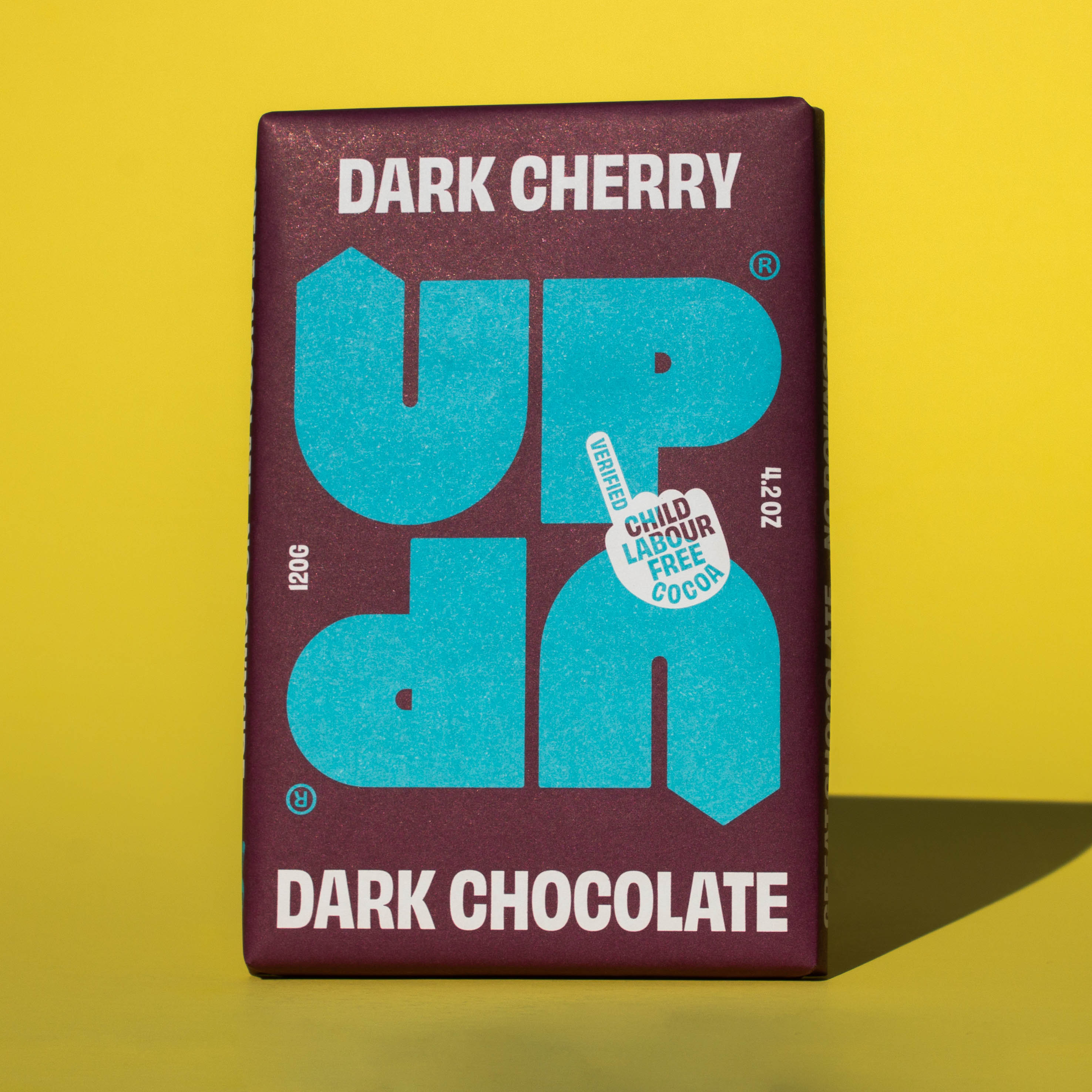 Bar of UP UP Dark Cherry Dark Chocolate, it has a purple paper wrapper with a blue logo, it is placed on a yellow background.