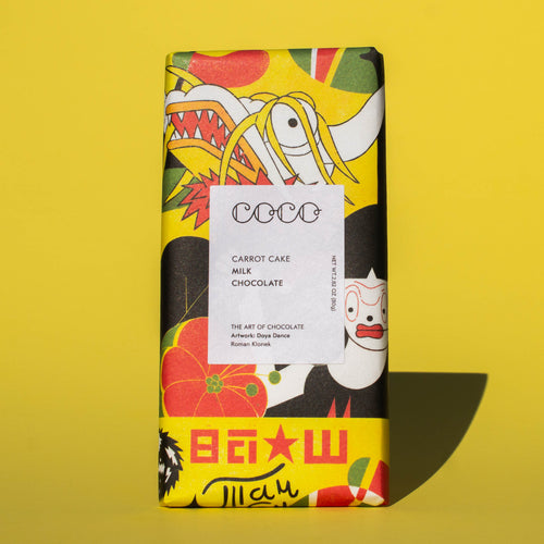 Bar of Coco Chocolatier Carrot Cake Milk Chocolate, it has a brightly colourful yellow, red and black illustration of a dragon on the wrapper, it is placed on a yellow background.