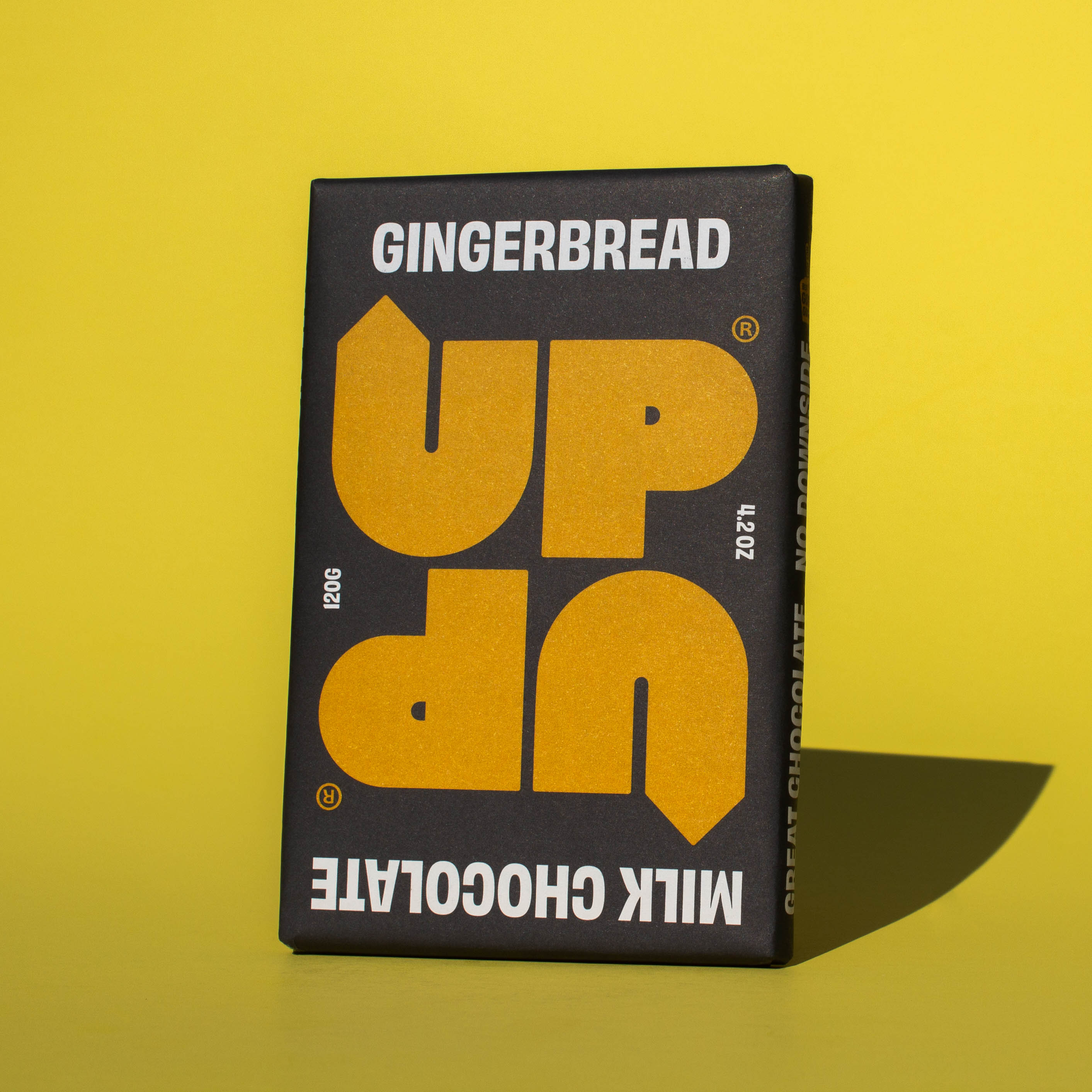 Bar of UP UP Gingerbread Milk Chocolate, it has a black paper wrapper with a yellow logo, it is placed on a yellow background and tilted slightly to the side.