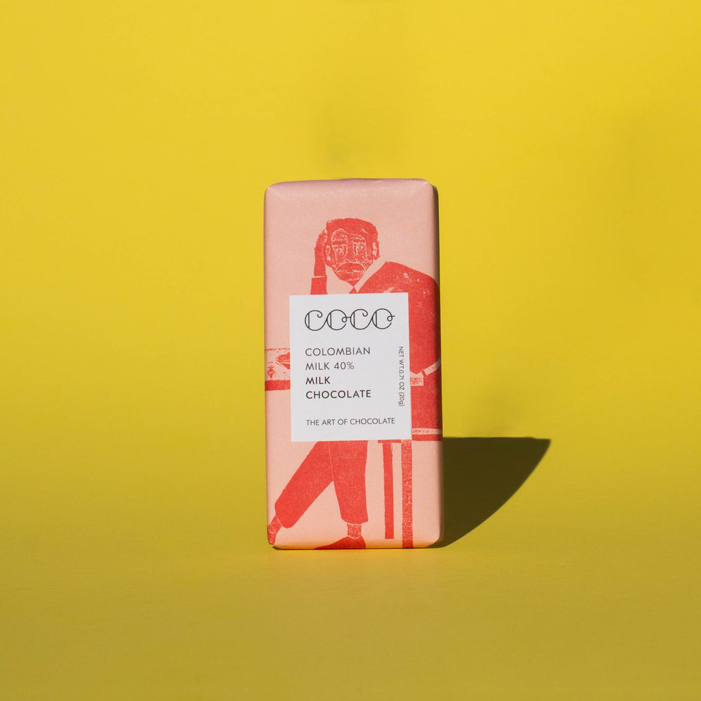 Bar of Coco Chocolatier Colombian Milk Chocolate, it has a pink wrapper with an illustration of a man sitting on a chair, it is placed on a yellow background.