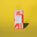 Bar of Coco Chocolatier Colombian Milk Chocolate, it has a pink wrapper with an illustration of a man sitting on a chair, it is placed on a yellow background.