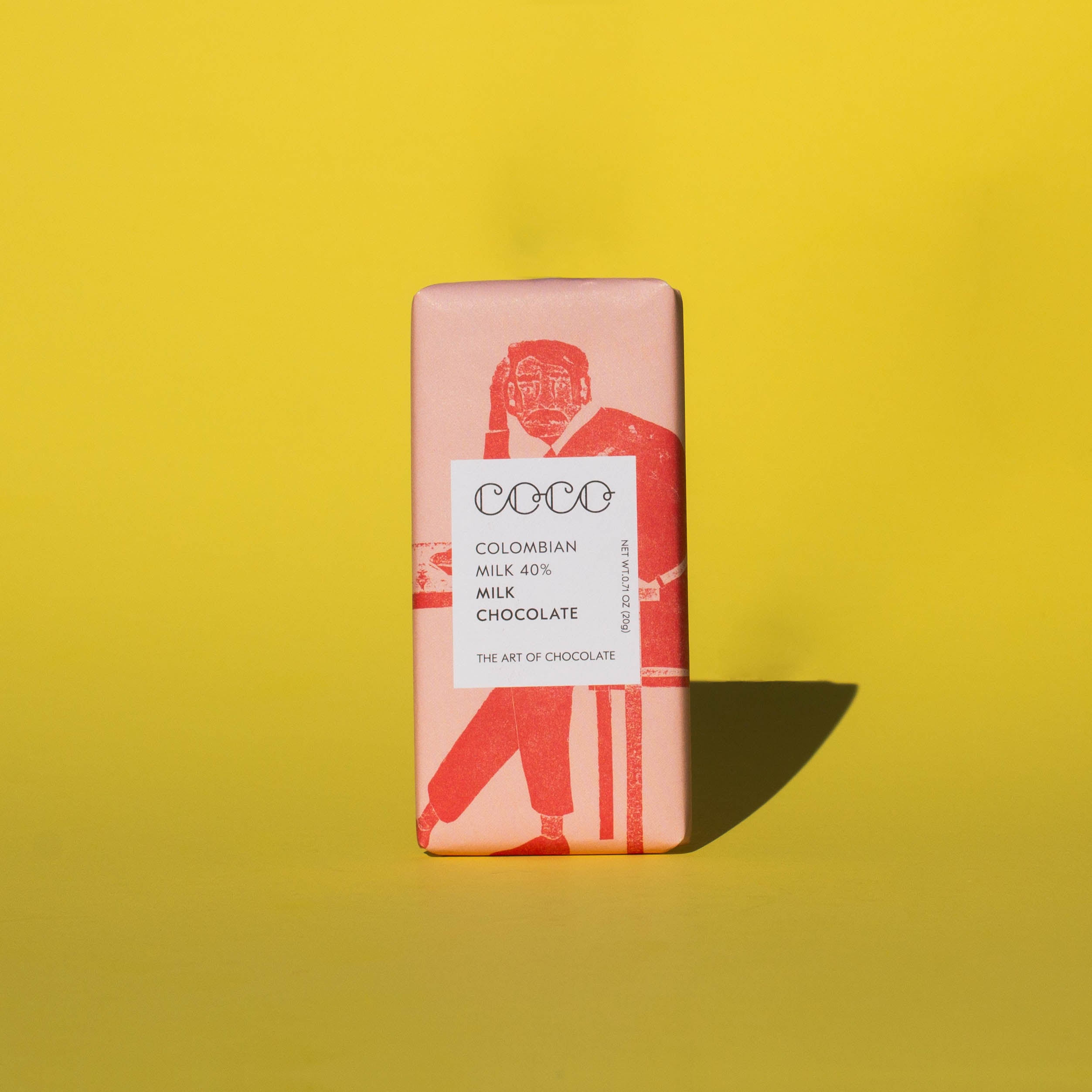 Bar of Coco Chocolatier Colombian Milk Chocolate, it has a pink wrapper with an illustration of a man sitting on a chair, it is placed on a yellow background.