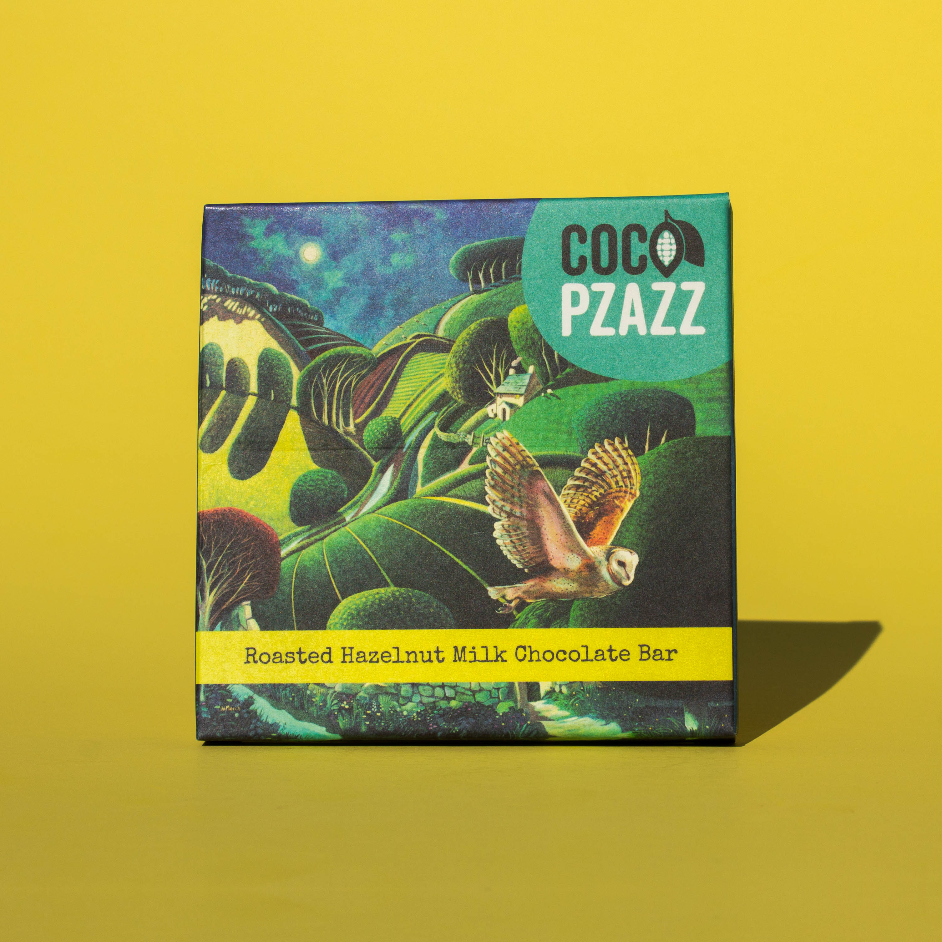 Bar of Coco Pzazz Roasted Hazelnut Milk chocolate, it has a dark blue wrapper with an illustration of an owl flying over farmland, it is placed on a yellow background.