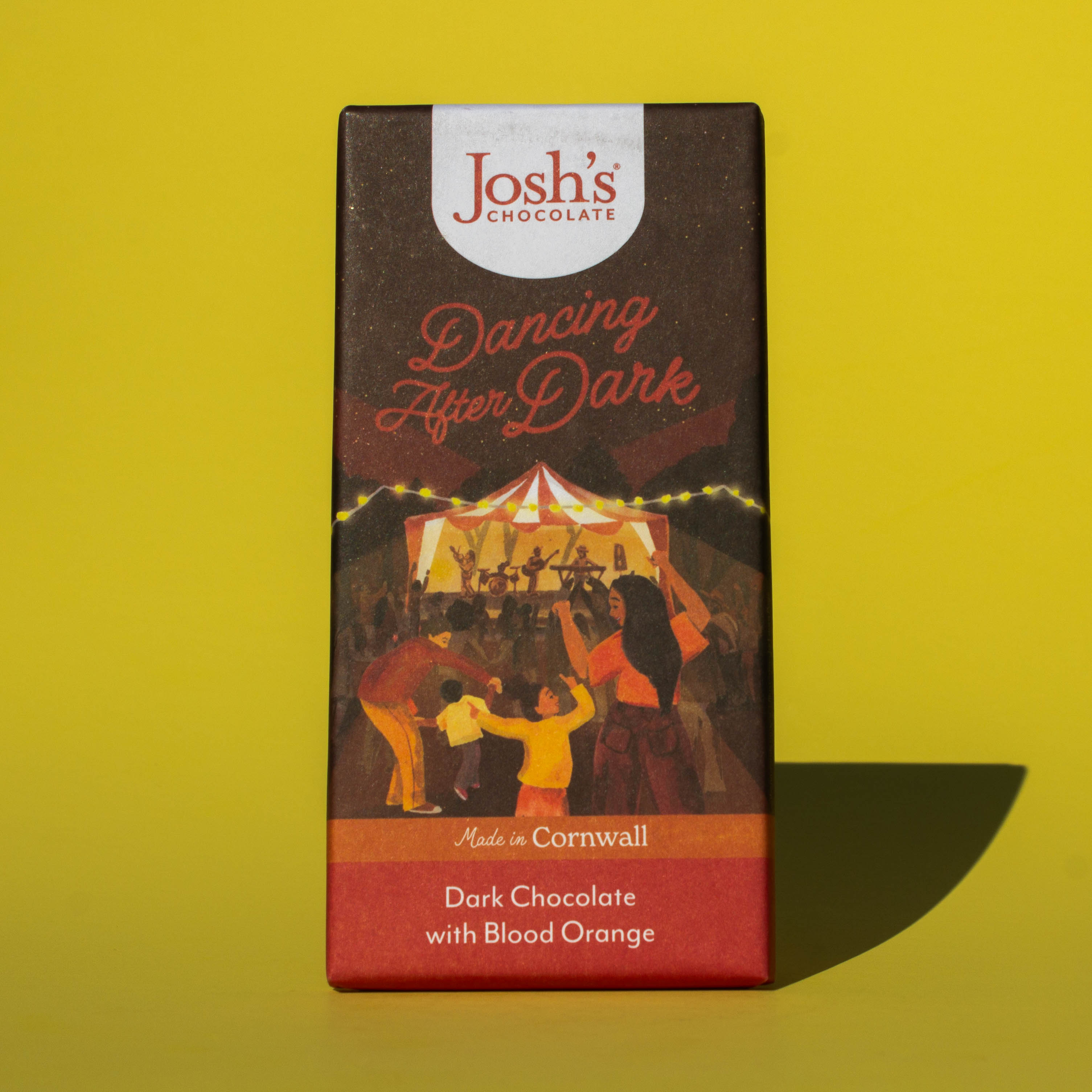 Bar of Josh's Dark Chocolate with Blood Orange, it is a dark orange and red paper wrapper with an illustration of an outdoor music event, it is placed on a yellow background.