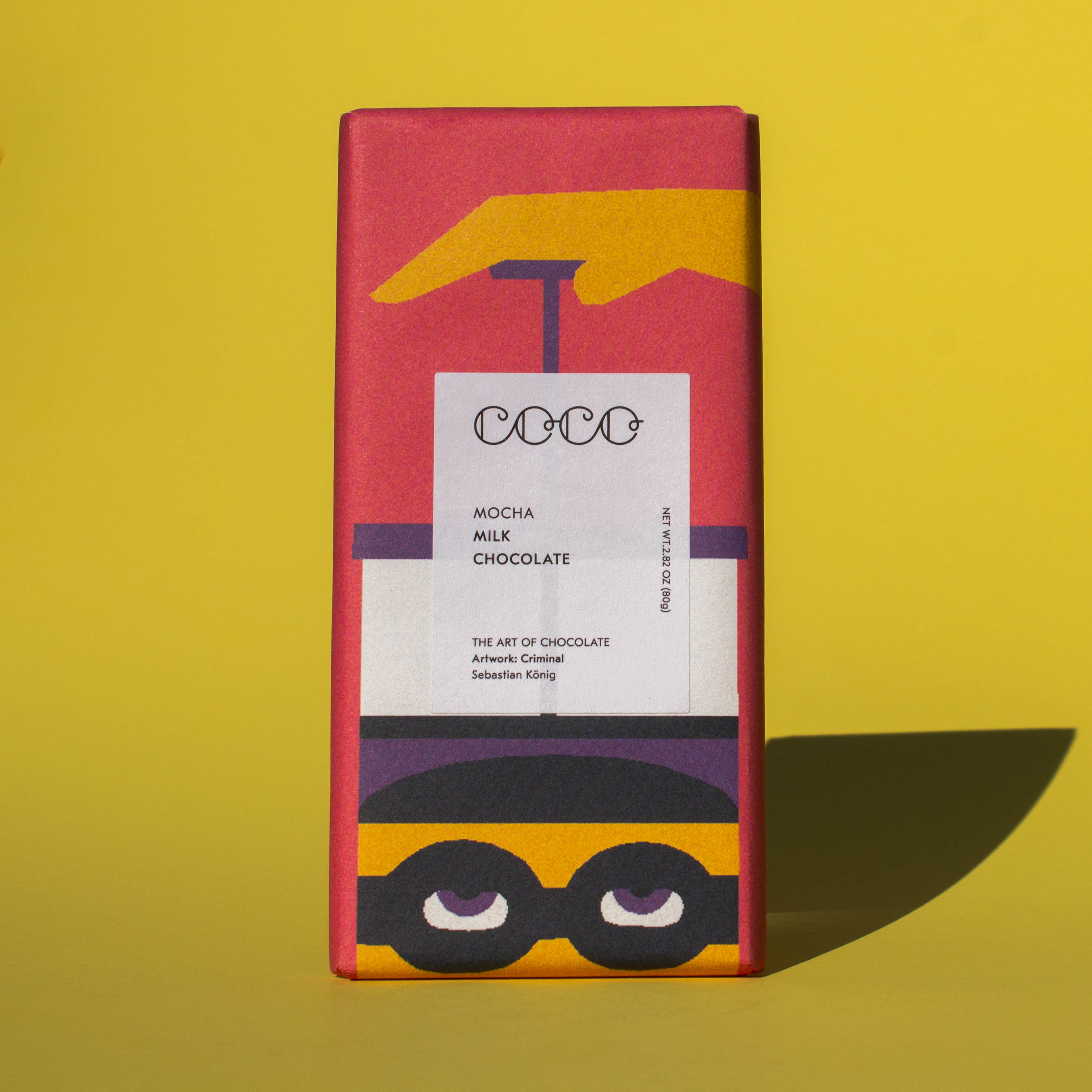 Bar of Coco Chocolatier Mocha Milk Chocolate, it has a pink wrapper with an illustration of a French press, it is placed on a yellow background.