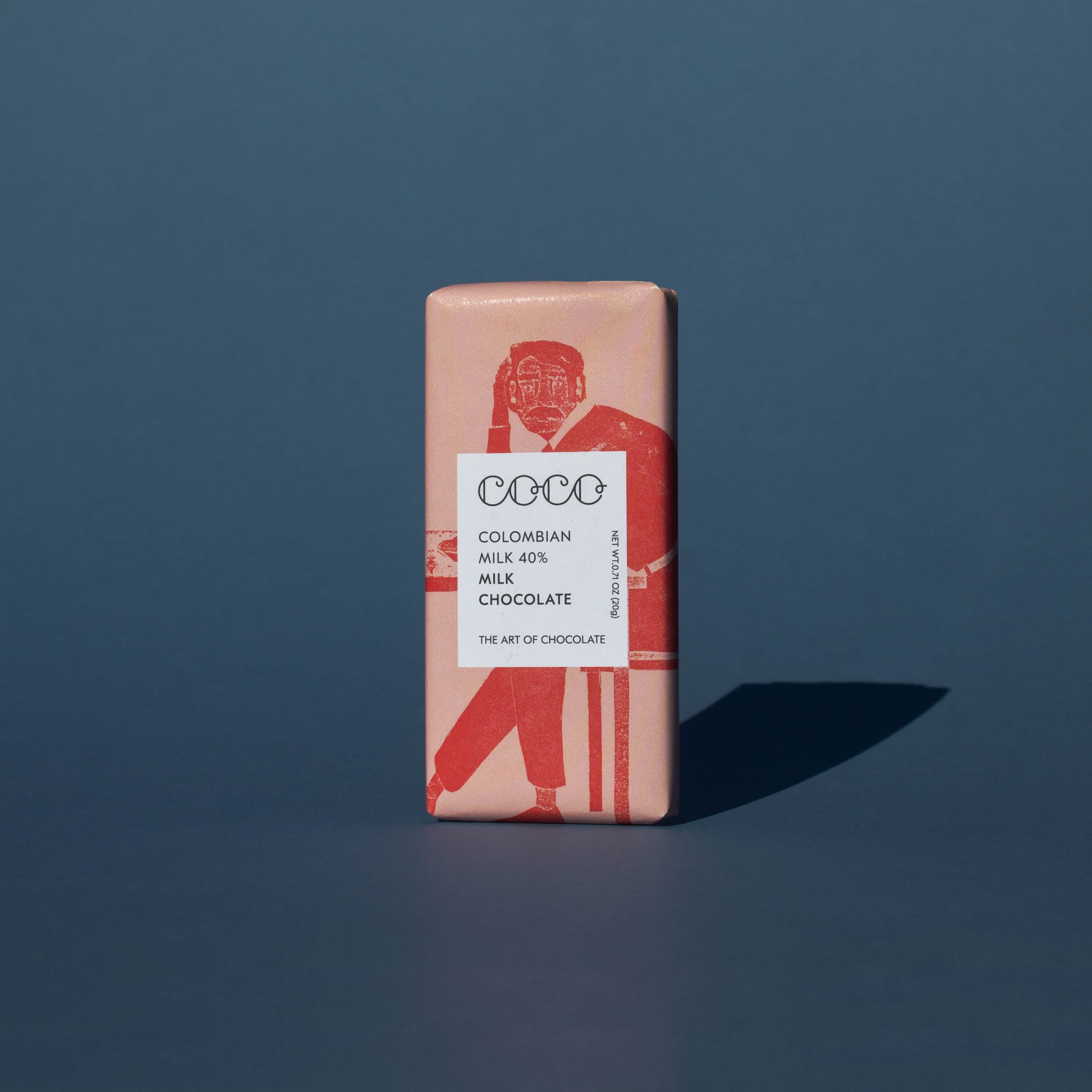 Bar of Coco Chocolatier Colombian Milk Chocolate, it has a pink wrapper with an illustration of a man sitting on a chair, it is placed on a blue background.