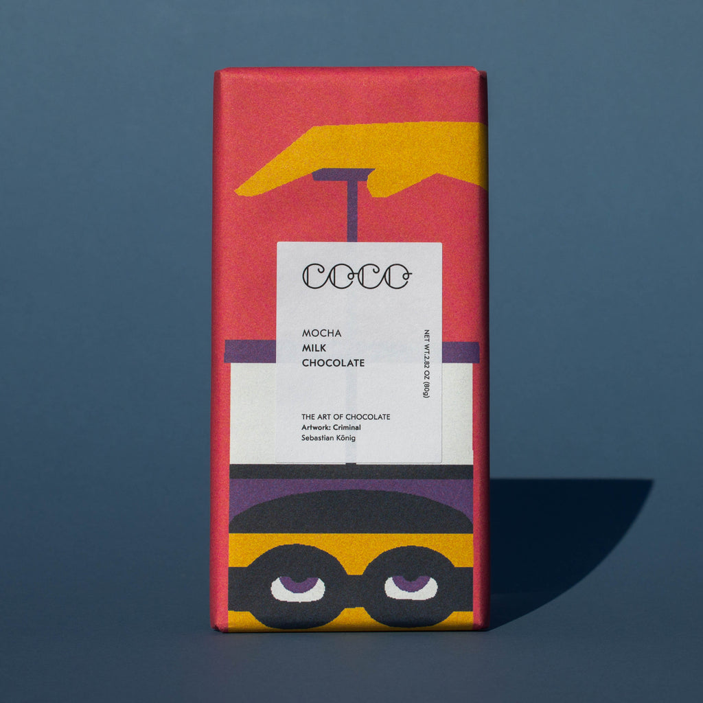 Bar of Coco Chocolatier Mocha Milk Chocolate, it has a pink wrapper with an illustration of a French press, it is placed on a blue background.