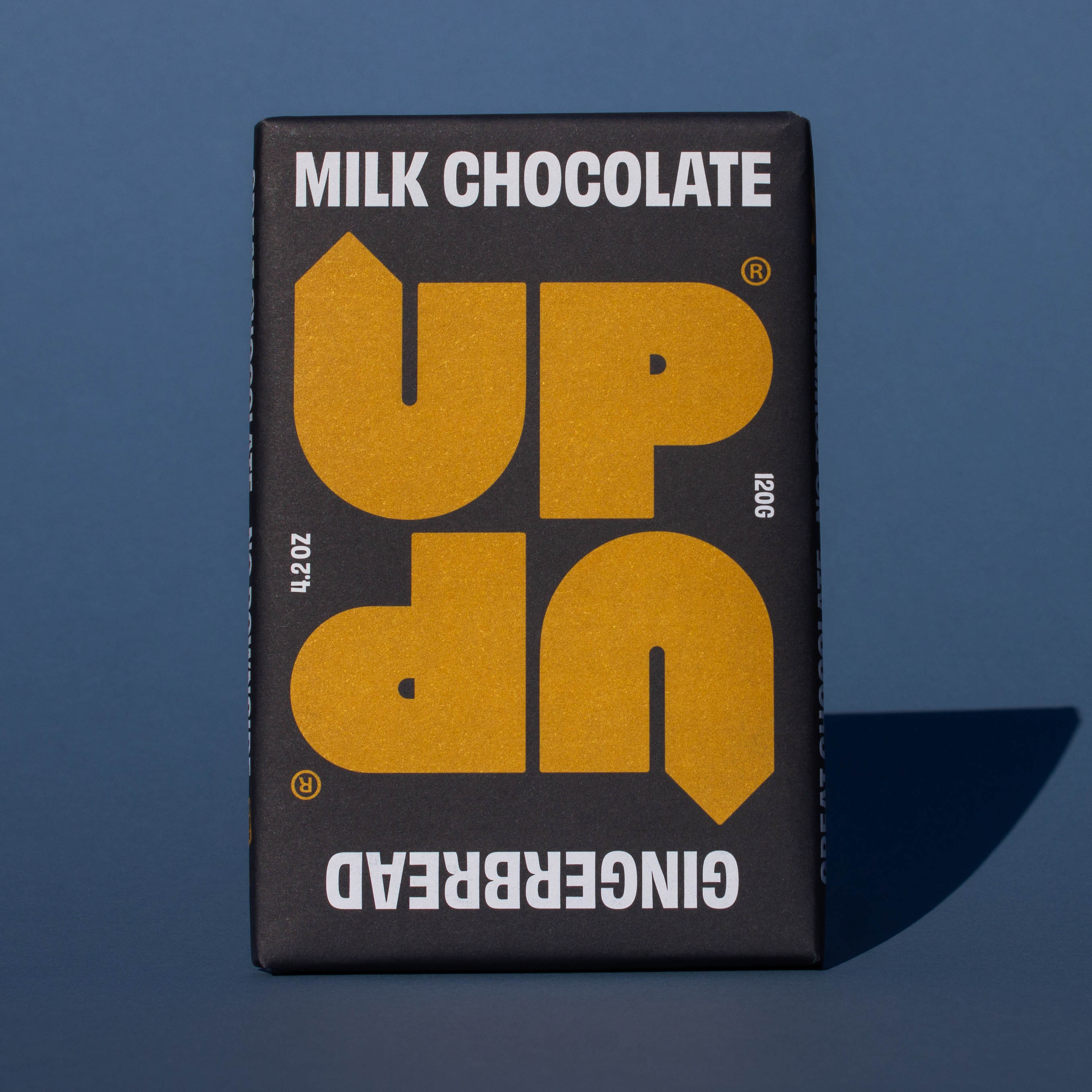 Bar of UP UP Gingerbread Milk Chocolate, it has a black paper wrapper with a yellow logo, it is placed on a blue background.