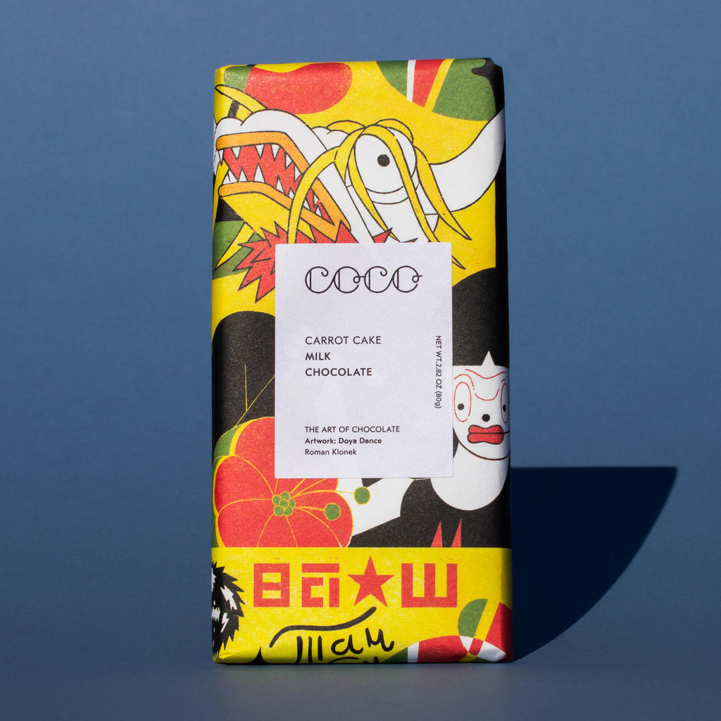 Bar of Coco Chocolatier Carrot Cake Milk Chocolate, it has a brightly colourful yellow, red and black illustration of a dragon on the wrapper, it is placed on a blue background.