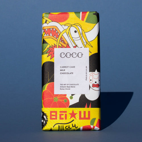 Bar of Coco Chocolatier Carrot Cake Milk Chocolate, it has a brightly colourful yellow, red and black illustration of a dragon on the wrapper, it is placed on a blue background.