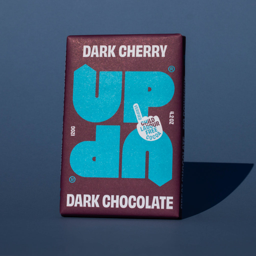 Bar of UP UP Dark Cherry Dark Chocolate, it has a purple paper wrapper with a blue logo, it is placed on a blue background and tilted slightly to the side.