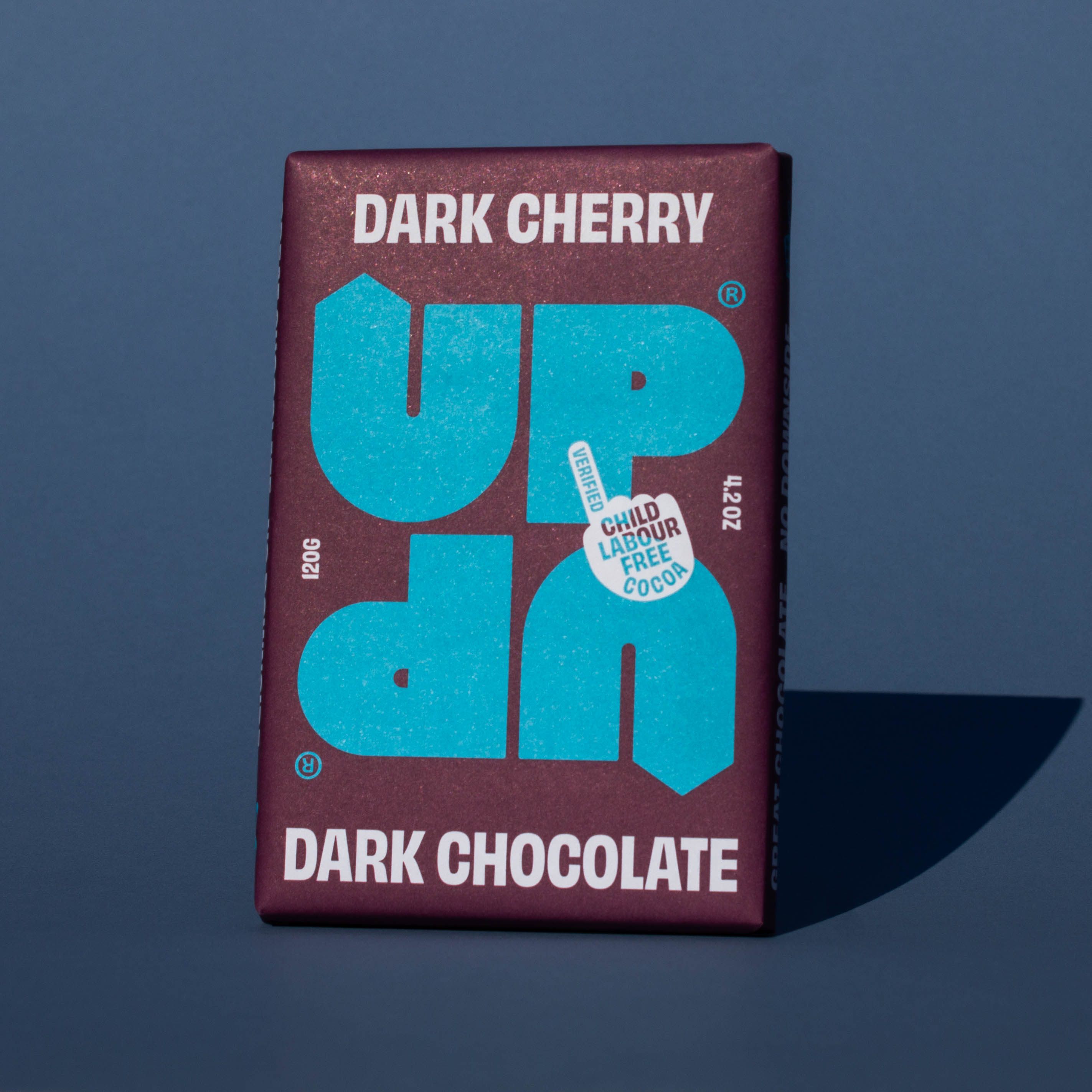 Bar of UP UP Dark Cherry Dark Chocolate, it has a purple paper wrapper with a blue logo, it is placed on a blue background and tilted slightly to the side.