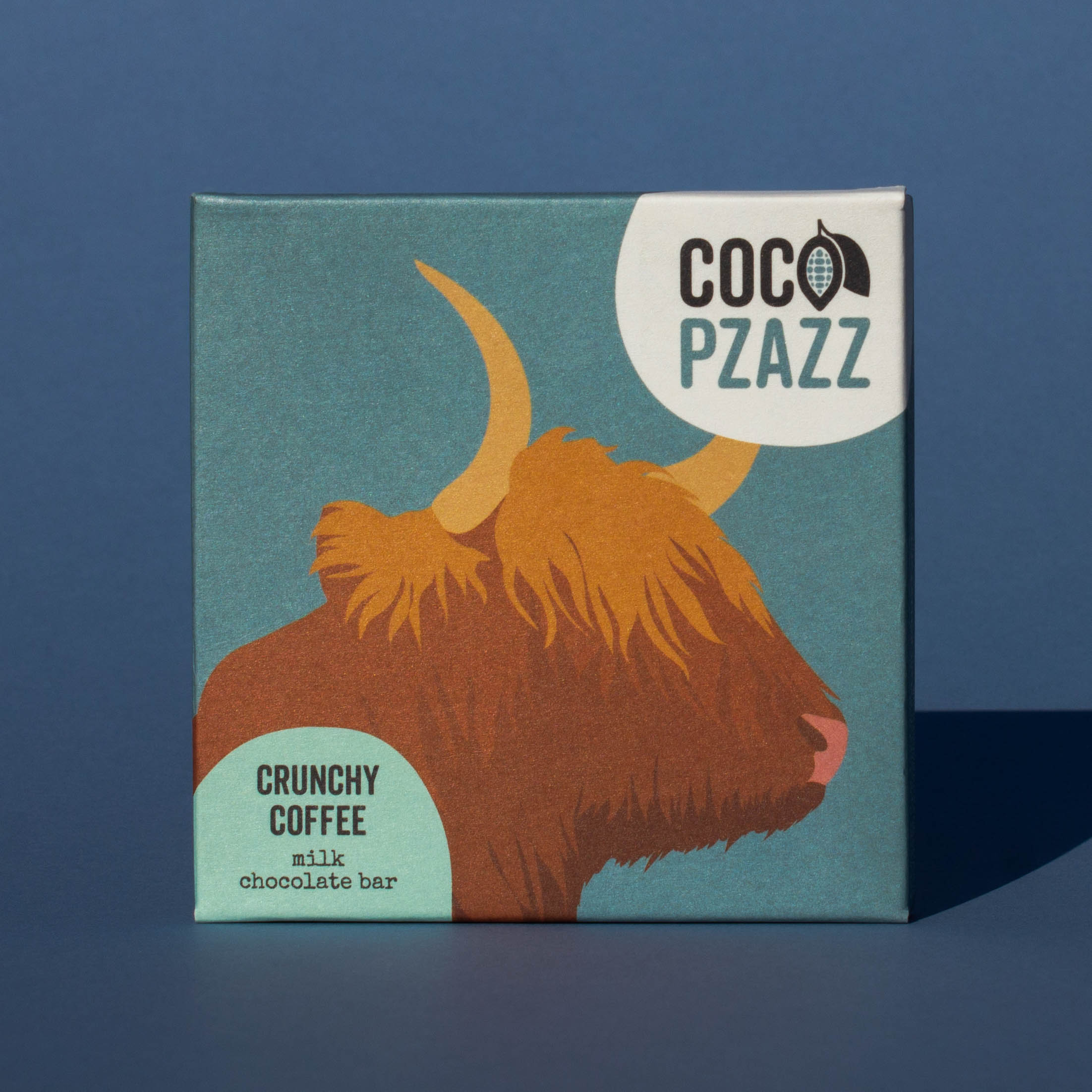Bar of Coco Pzazz Crunchy Coffee Milk Chocolate, it has a blue wrapper with an illustration of a highland cow, it is placed on a blue background.