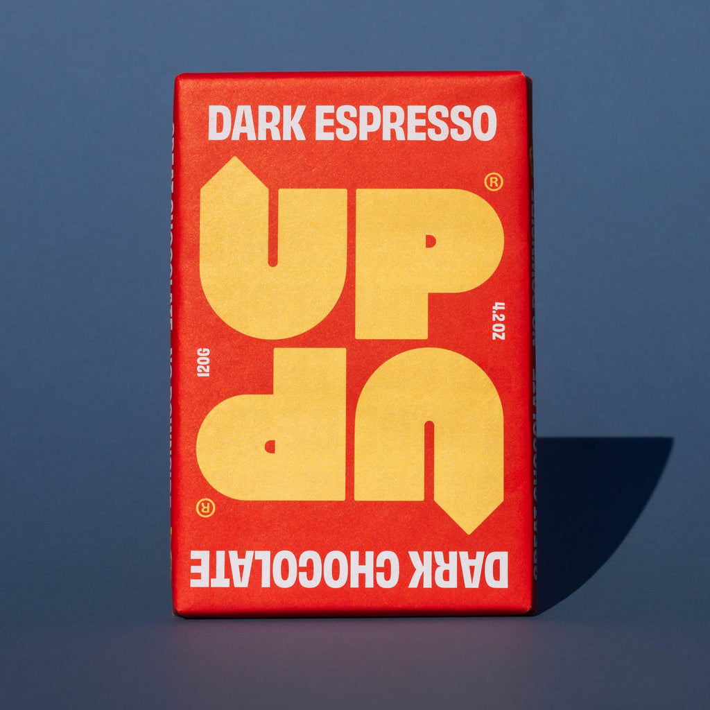Bar of UP UP Dark Espresso Dark Chocolate, it has a red paper wrapper with a yellow logo, it is placed on a blue background.