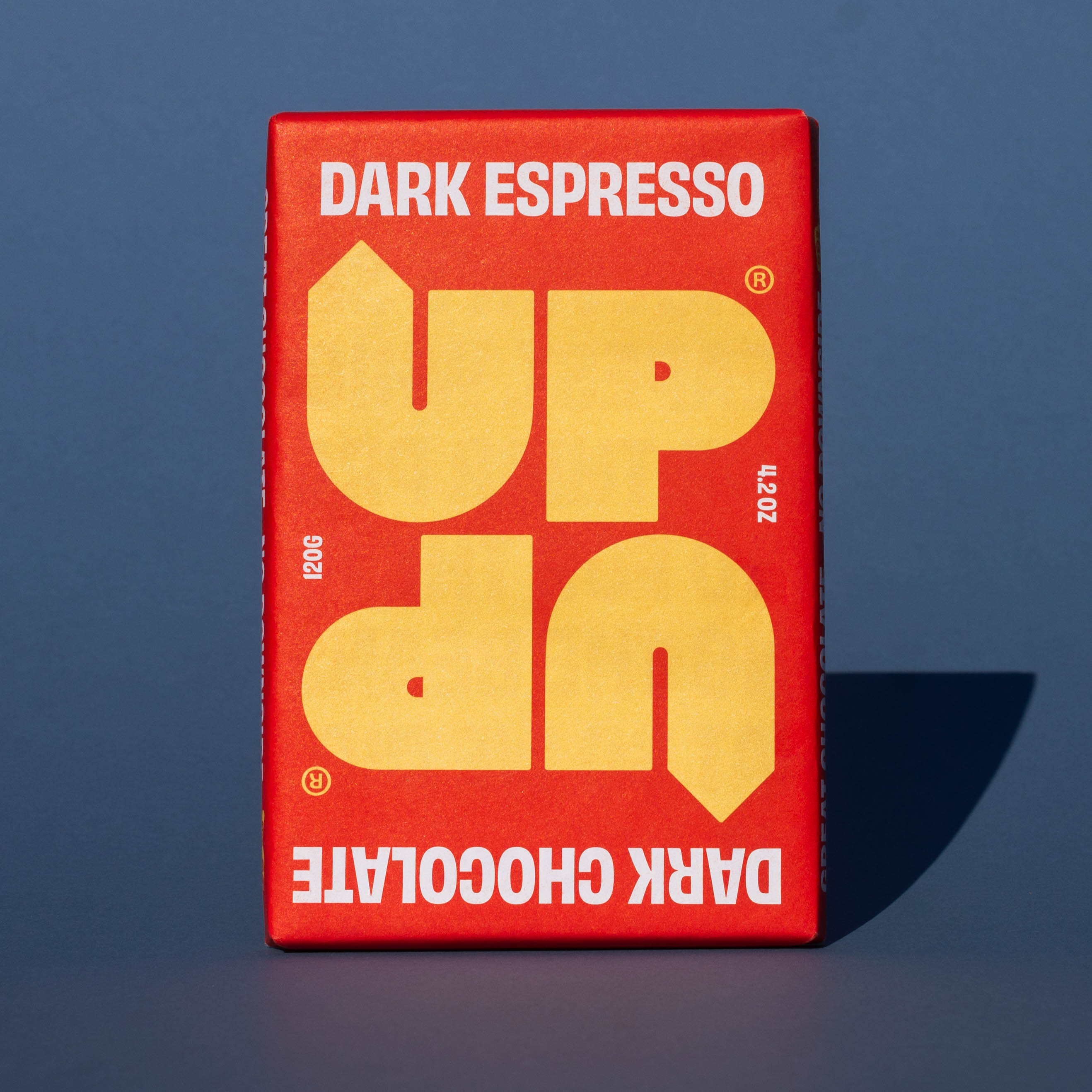 Bar of UP UP Dark Espresso Dark Chocolate, it has a red paper wrapper with a yellow logo, it is placed on a blue background.