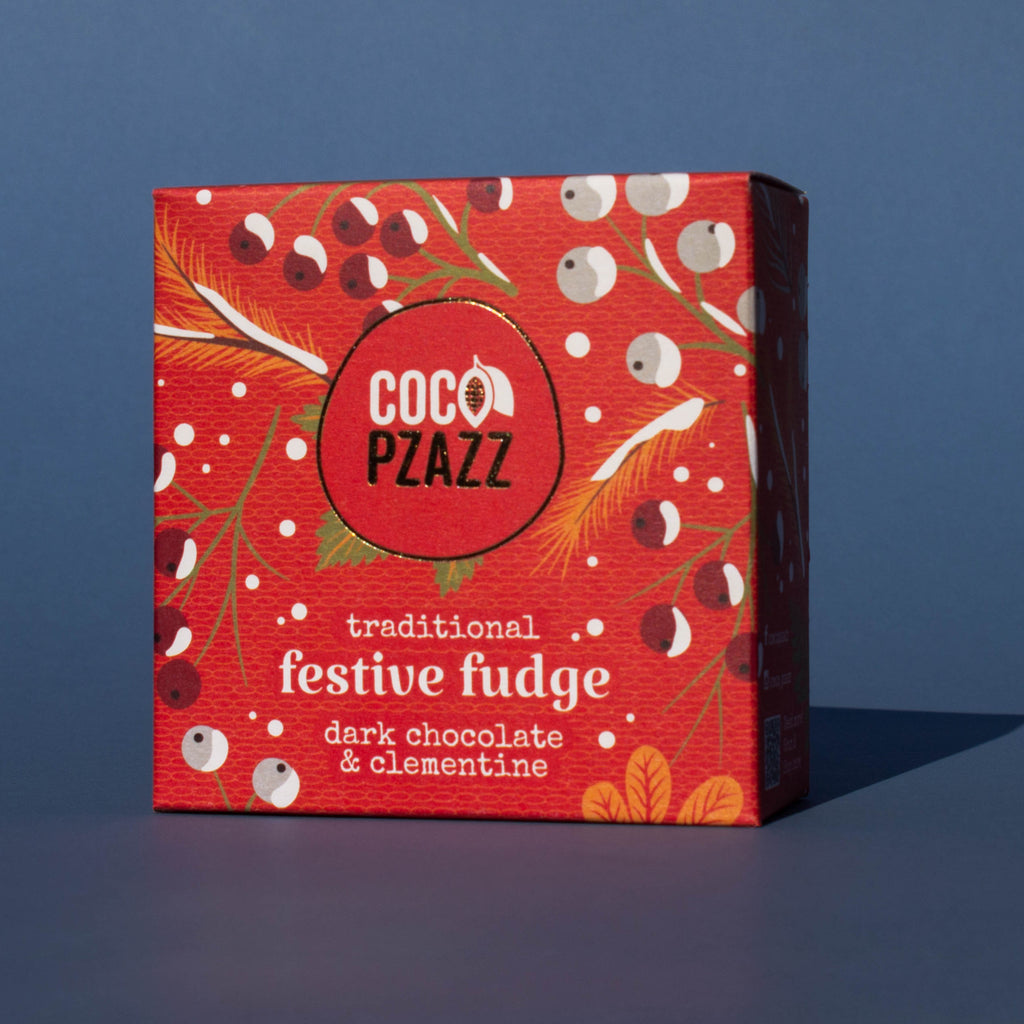 Box of Coco Pzazz Dark Chocolate and Clementine Fudge, it is a red box with an illustration of winter berries, it is placed on a blue background and tilted slightly to the side.