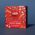 Box of Coco Pzazz Dark Chocolate and Clementine Fudge, it is a red box with an illustration of winter berries, it is placed on a blue background and tilted slightly to the side.