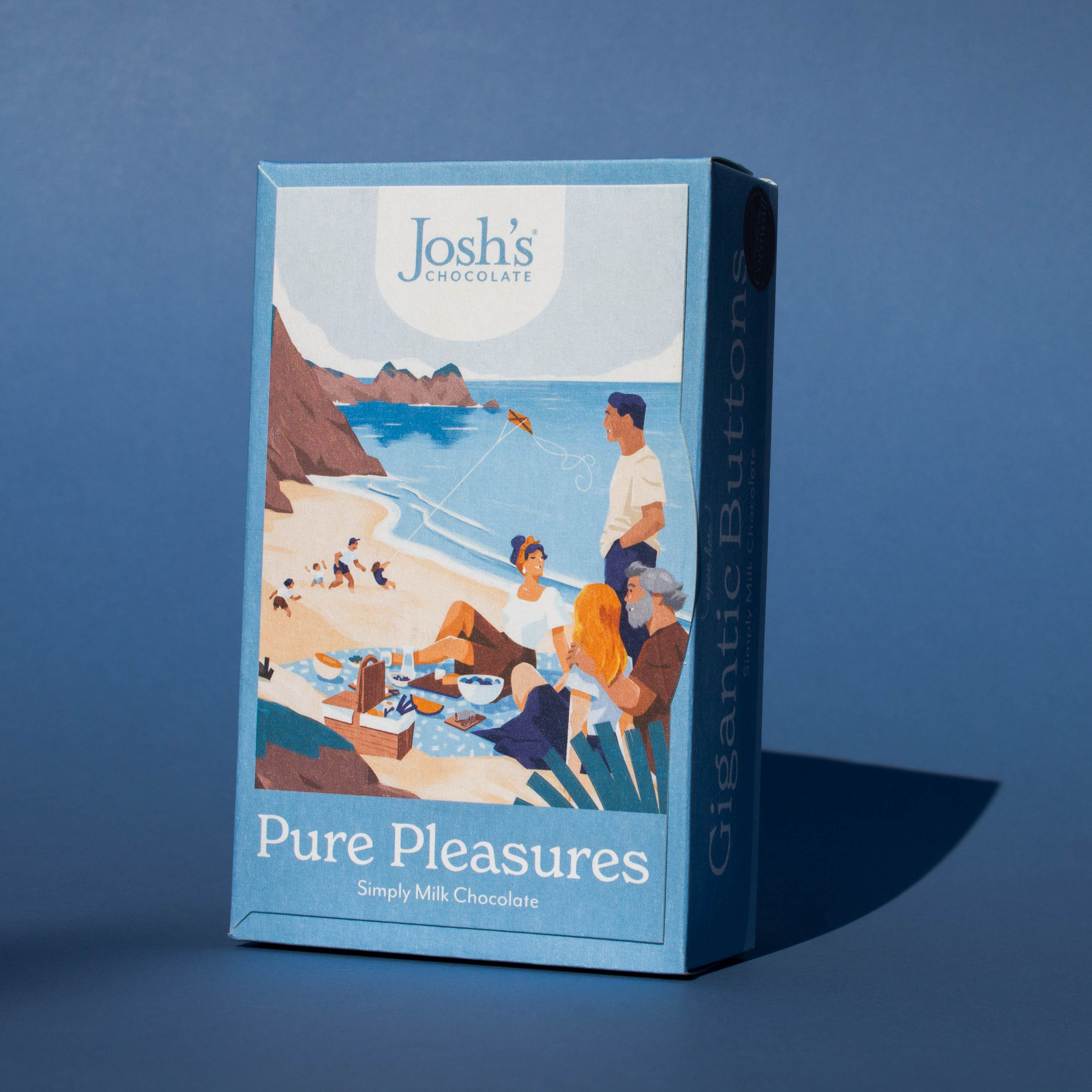 Box of Josh's Milk Chocolate Buttons tilted to the side, it is a blue box an with illustration of a beach picnic scene, it is placed on a blue background.