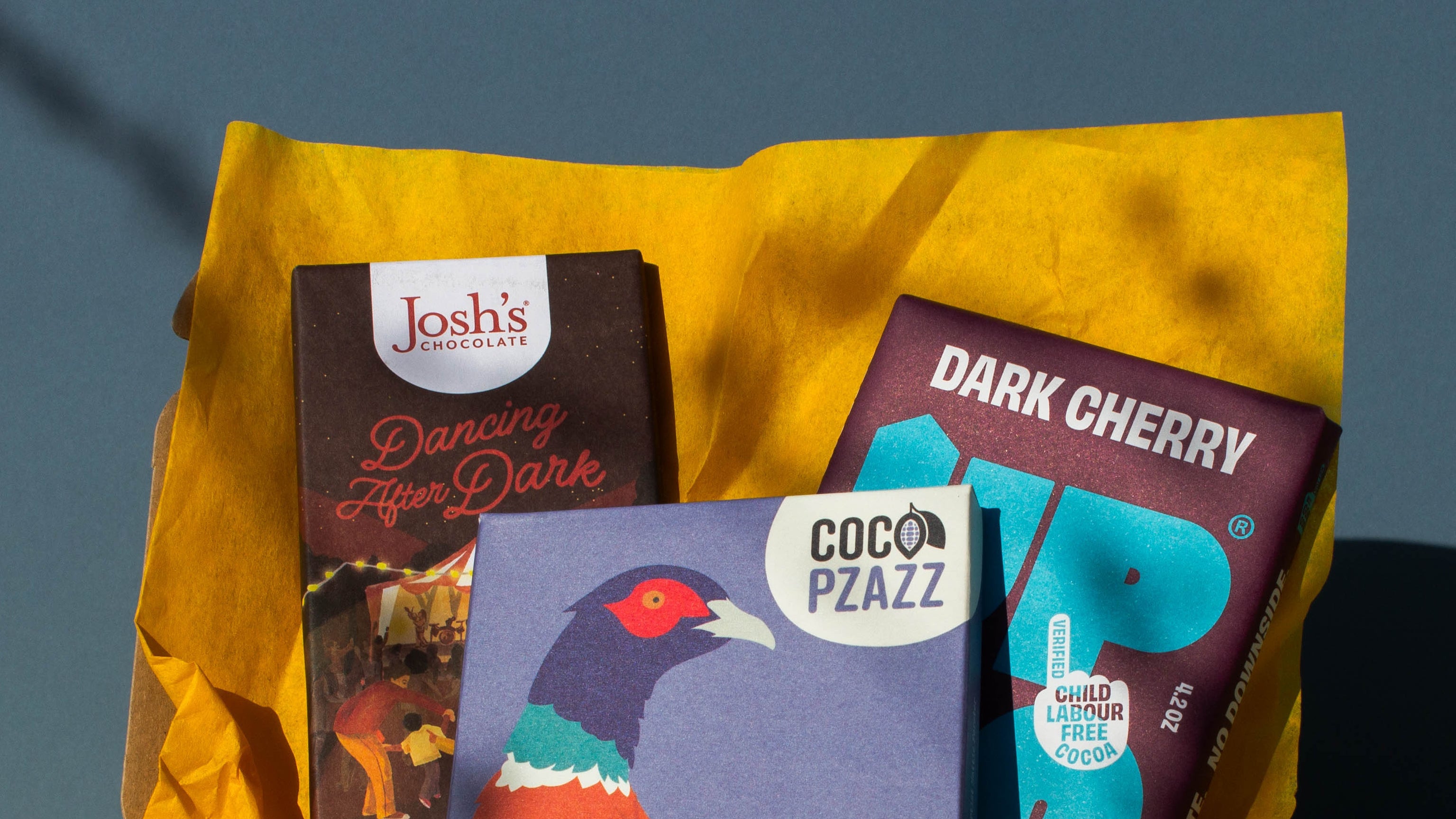Top of Gift box filled with yellow wrapping paper and holding 3 bars of chocolate, Josh's Blood Orange Dark Chocolate Up Up Dark Cherry, and Coco Pzazz Ecuador Dark Chocolate, it is cast over with shadow, and is on a blue background.