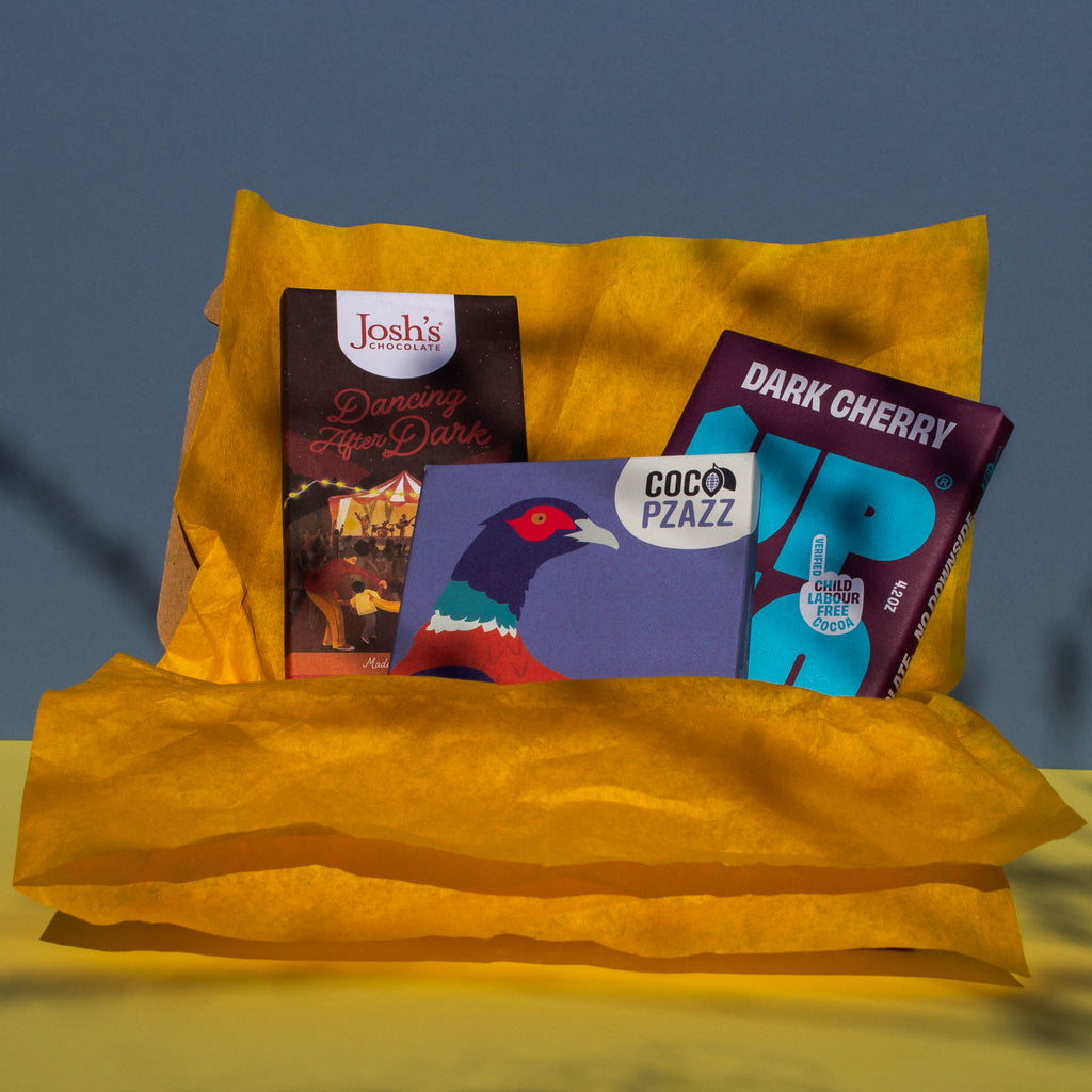 Gift box filled with yellow wrapping paper and holding 3 bars of chocolate, Josh's Blood Orange Dark Chocolate Up Up Dark Cherry, and Coco Pzazz Ecuador Dark Chocolate, it is cast over with shadow, and is on a yellow and blue background.
