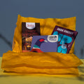 Gift box filled with yellow wrapping paper and holding 3 bars of chocolate, Josh's Blood Orange Dark Chocolate Up Up Dark Cherry, and Coco Pzazz Ecuador Dark Chocolate, it is cast over with shadow, and is on a yellow and blue background.