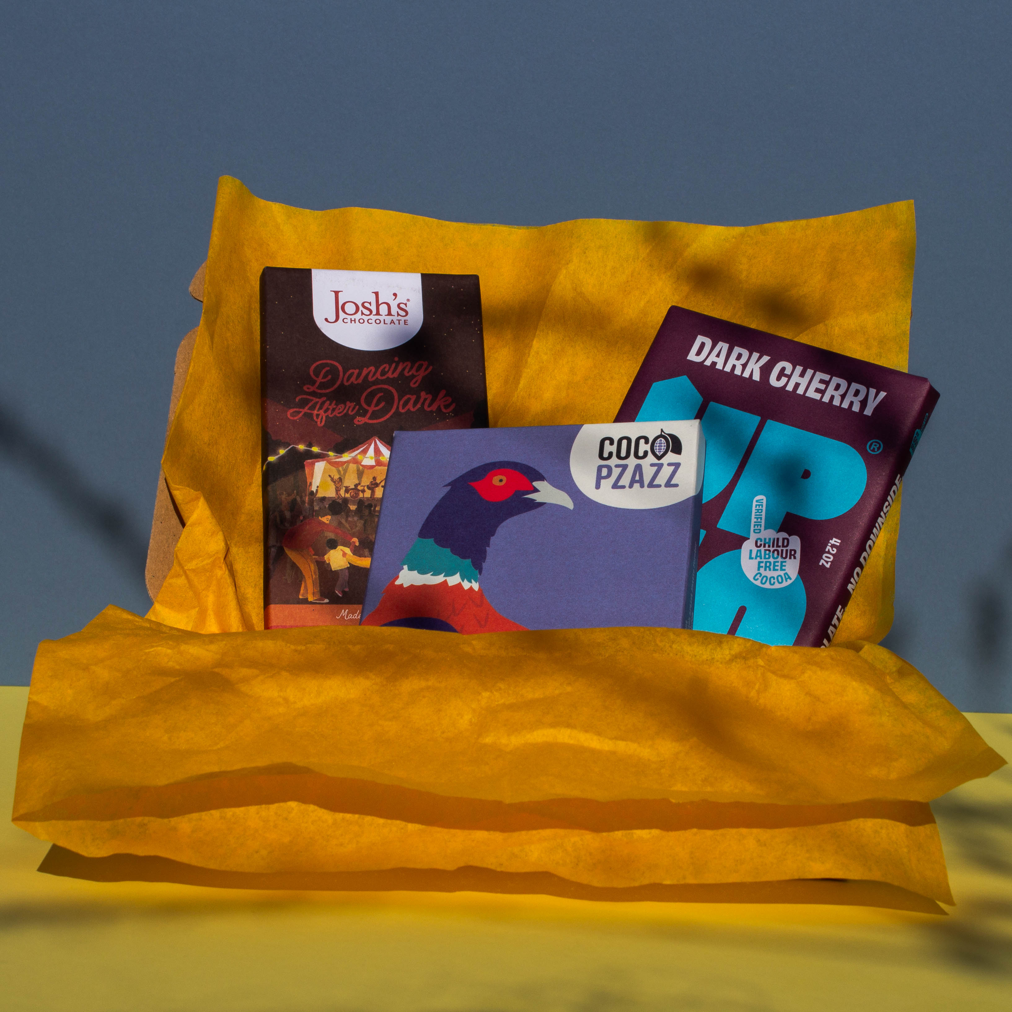 Gift box filled with yellow wrapping paper and holding 3 bars of chocolate, Josh's Blood Orange Dark Chocolate Up Up Dark Cherry, and Coco Pzazz Ecuador Dark Chocolate, it is cast over with shadow, and is on a yellow and blue background.