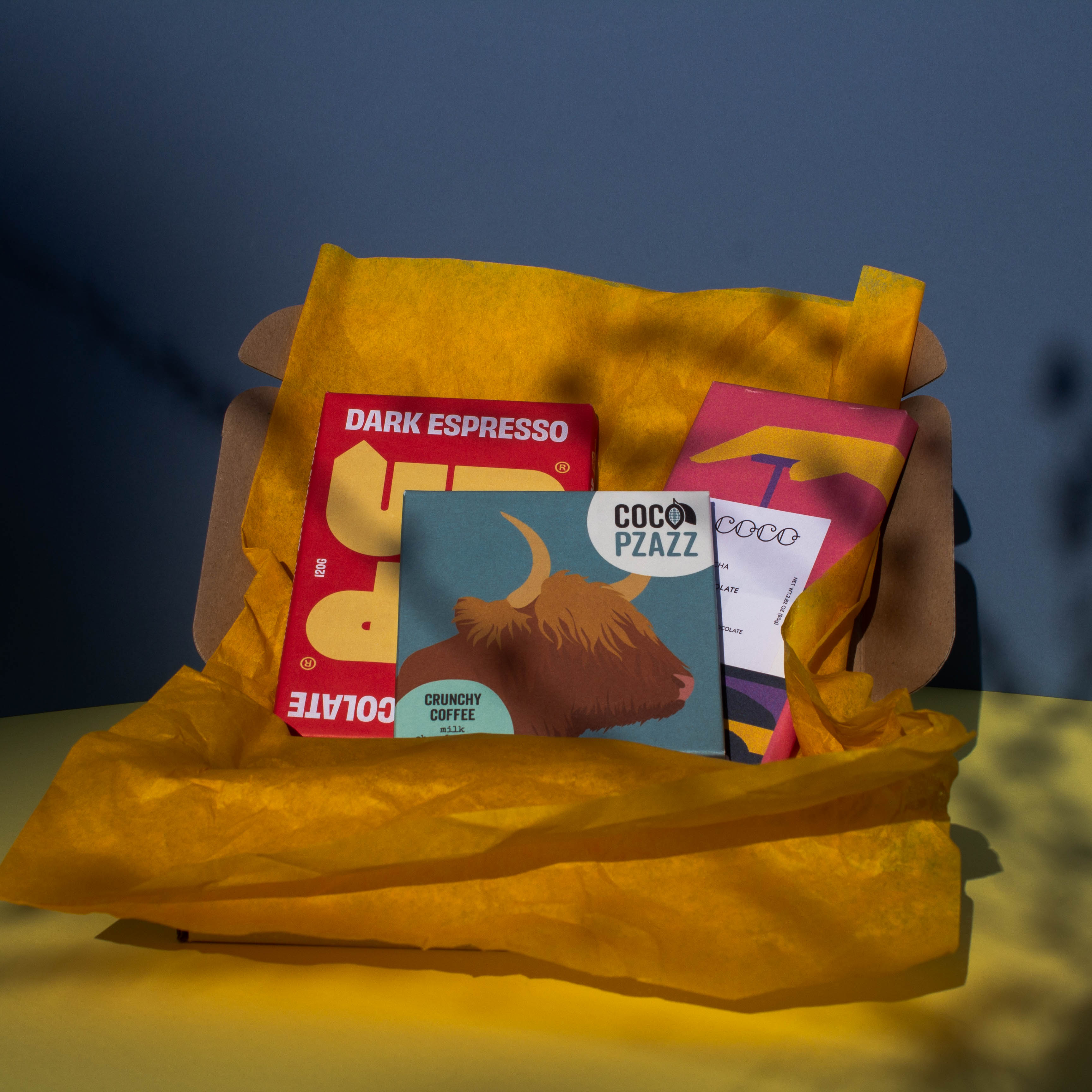 Gift box filled with yellow wrapping paper and holding 3 bars of chocolate, Up Up Dark Espresso, Coco Pzazz Crunchy Coffee and Coco Chocolatier Mocha Milk, it is cast over with shadow, and is on a yellow and blue background.