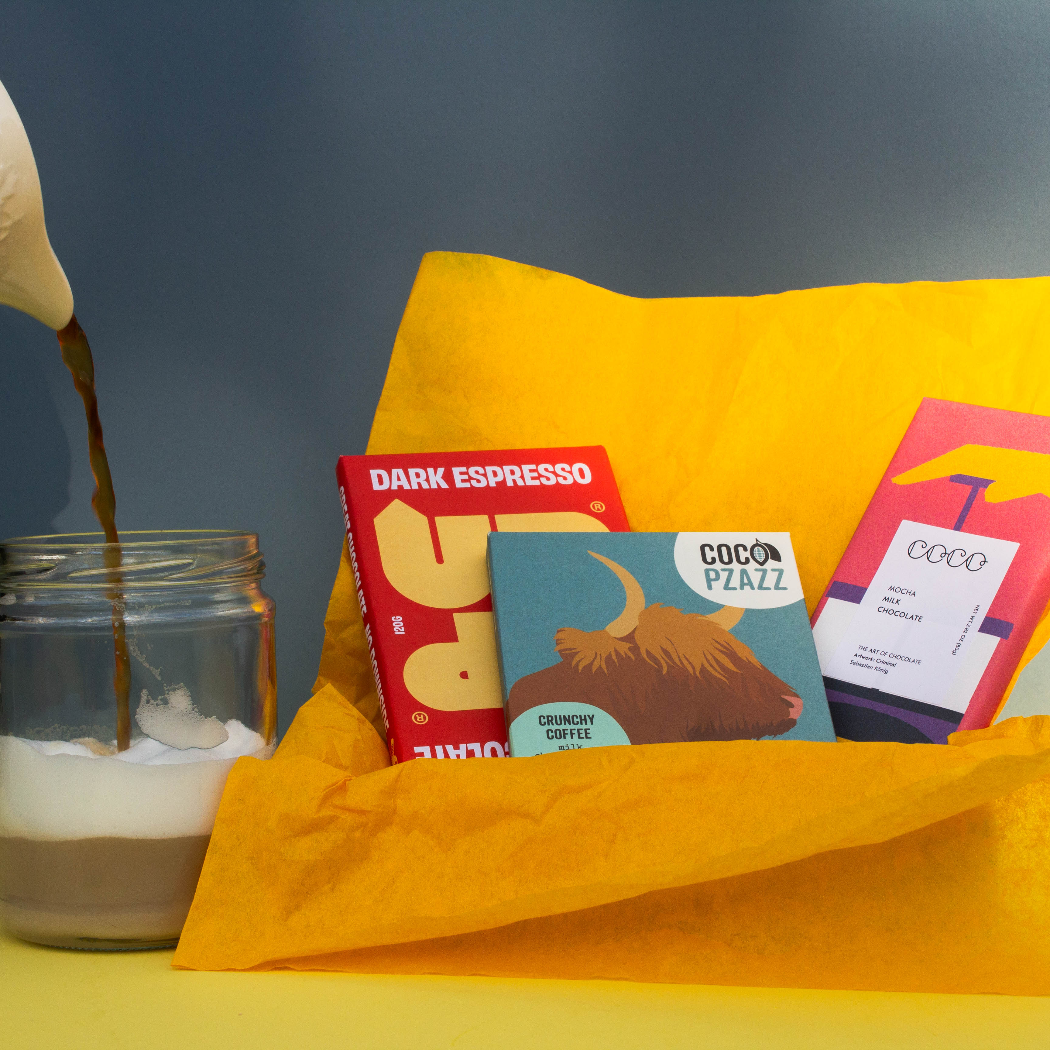 Gift box filled with yellow wrapping paper and holding 3 bars of chocolate, Up Up Dark Espresso, Coco Pzazz Crunchy Coffee and Coco Chocolatier Mocha Milk. Next to it is a jug pouring coffee into a jar with foamy coffee inside of it. Both are on a yellow and blue background.